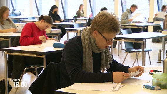 Schüler*innen sitzen an Tischen in einer Halle © Screenshot 