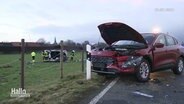 Nach einem Verkehrsunfall auf einer Landstraße steht ein rotes Auto mit Totalschaden und aufgeklappter Motohaube am Straßenrand, auf einem nahegelegenen Feld stehen Rettungskräfte bei einem auf der Seite liegendem Auto. © Screenshot 