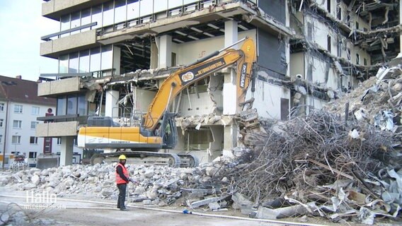 Ein Bauarbeiter mit orangefarbener Warnweste steht vor einer größeren Abrissbaustelle eines Hochhauses mit einem größeren Abrissbagger. © Screenshot 