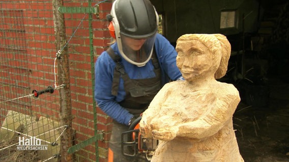 Eine Frau mit Arbeitsschutzkleidung bearbeitet mit einer Kettensäge eine Holzfigut einer Frau, die ihre Arme nach vorne ausstreckt. © Screenshot 