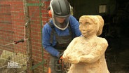 Eine Frau mit Arbeitsschutzkleidung bearbeitet mit einer Kettensäge eine Holzfigut einer Frau, die ihre Arme nach vorne ausstreckt. © Screenshot 