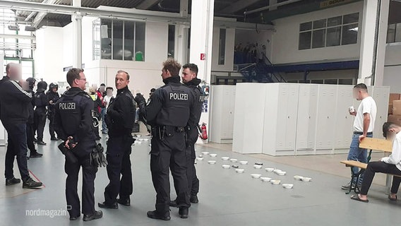 In einer größeren Halle stehen mehrere Einsatzkräfte der Polizei, dahinter stehen mehrere Schüsseln aufgereiht auf dem Boden. © Screenshot 