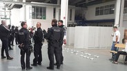 In einer größeren Halle stehen mehrere Einsatzkräfte der Polizei, dahinter stehen mehrere Schüsseln aufgereiht auf dem Boden. © Screenshot 