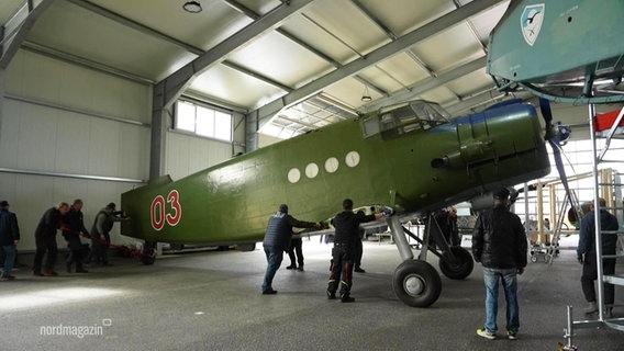 Mehrere Personen schieben ein größeres Doppeldecker-Propellerflugzeug in einer Hangar-Halle. © Screenshot 