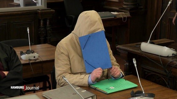 Ein Angeklagter im beigen Kapuzenpulli hält sich eine blaue Mappe vor das Gesicht auf der Anklagebank. © Screenshot 