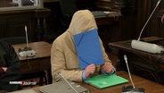 Ein Angeklagter im beigen Kapuzenpulli hält sich eine blaue Mappe vor das Gesicht auf der Anklagebank. © Screenshot 