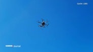 Blick aus der Froschperspektive auf eine kleinere Drohne die vor einem blauen Himmel fliegt. © Screenshot 