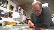 Ein Mann malt mit einer Spritzdose Farbe auf ein größeres Bild in einem Atelier. © Screenshot 
