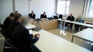 In einem Verhandlungssaal sitzen Kläger und Angeklagte sowie die Richter beieinander. © Screenshot 