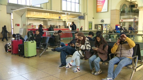 Wartende Menschen sitzen in einer Bahnhofshalle. © Screenshot 