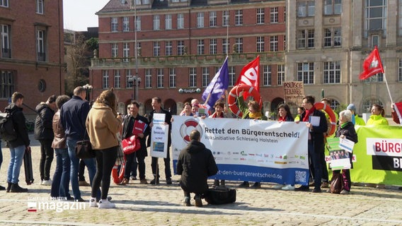 Demonstrierende Menschen stehen mit Plakaten für ein Foto zusammen. © Screenshot 