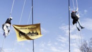 Zwei Menschen hängen an Seilen mit einem Banner in der Hand © Screenshot 
