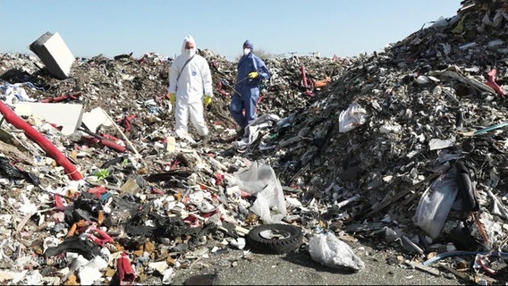 Zwei Menschen in Schutzkleidung stehen zwischen Bergen von Müll. © Screenshot 