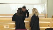Ein Angeklagter steht mit seinen Anwältinnen im Gerichtssaal. © Screenshot 