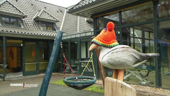 Blick auf den Innenhof eines Kinderhospiz: Eine Holzmöwe mit Mütze sitzt auf einem Pfahl, im Hintergrund eine Korbschaukel. © Screenshot 