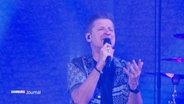 Der Sänger von PUR, Hartmut Engler, singt auf einer Bühne vor blauem Hintergrund bei einem Auftritt. © Screenshot 
