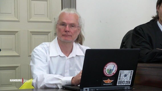 Ein Mann mit längeren grauen Haaren sitzt in einem Gerichtssaal. Vor sich hat er einen Laptop aufgeklappt. © Screenshot 
