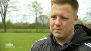 Ein jüngerer Mann in Trainingsjacke blickt auf einem Fußballplatz an der Kamera vorbei. © Screenshot 