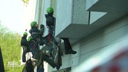 An einer Fassade hängen mehrere Aktivisten mit grünen Helmen, die sich abgeseilt haben. © Screenshot 