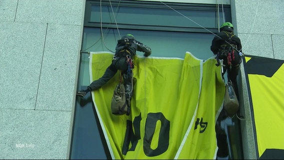 Zwei Aktivisten von Greenpeace montieren in Kletterseilen hängend ein größeres Banner an einer Fassade. © Screenshot 