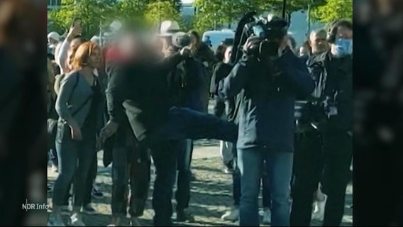 Ein Mann mit unkenntlich gemachtem Gesicht tritt einem Kameramann vor einer Menschentraube ins Gesäß. © Screenshot 
