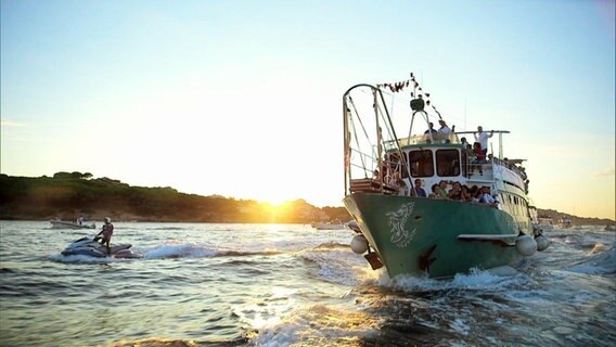 Ein kleines Fischerboot voller feiernden Menschen, unterwegs vor der Küste Sardiniens. © Screenshot 
