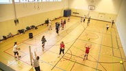 Blick in eine Halle, in der ein Volleyballnetz quer durch gespannt wurde, damit zahlreiche Kinder zusammen spielen können. © Screenshot 