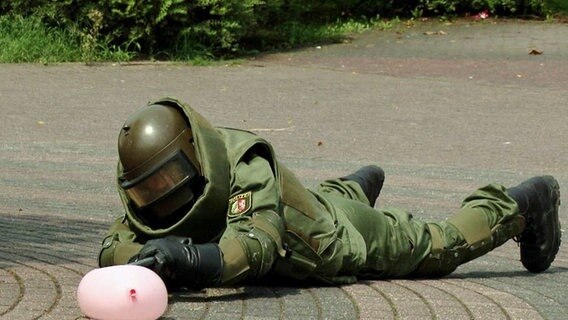 Ein Bombenentschärfer in Schutzkleidung liegt auf der Straße und macht sich an einer Wasserbombe zu schaffen.  