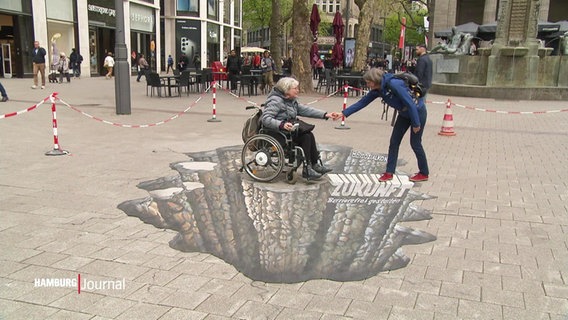 Eine Frau zieht eine andere Frau im Rollstuhl von einem Bodengemälde eines Abgrunds in einer Fußgängerzone. © Screenshot 