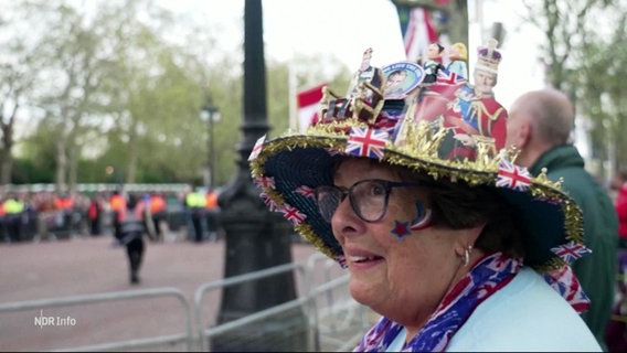 Eine Zuschauerin mit einem bunten Hut voller britischer Flaggen und Accessoires guckt sich belustigt die Straße vor dem Buckingham Palace an. © Screenshot 
