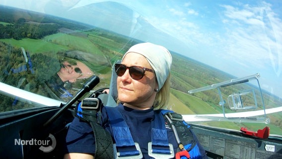 Eine Segelfliegerin sitzt im Cockpit eines kleinen Flugzeuges. Hinter ihr ist die Landschaft zusehen, über die sie fliegt. © Screenshot 