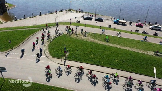 Viele Radfahrer fahren eine Straße entlang in Richtung eines kleinen Fähranlegers. Das Bild ist aus der Luft aufgenommen. © Screenshot 