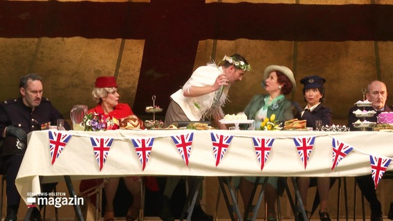 Mehrere Schauspielerinnen und Schauspieler sitzen auf der Bühne hinter einem langen Tisch, der mit englischen Fahnen geschmückt ist. © Screenshot 
