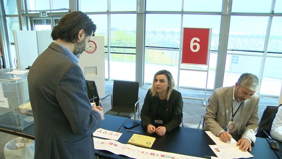 Selman Öz von der türkischen Regierungspartei AKP steht vor einem Wahlstand am Messegelände in Hannover. © Screenshot 