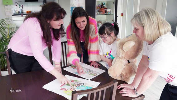 Die Autorinnen Annabell Behrmann und Miriam Opresmik, Valentina und Trainerin Maike stehen um Bilder aus einem Bilderbuch, das Valentinas Geschichte erzählt. © Screenshot 