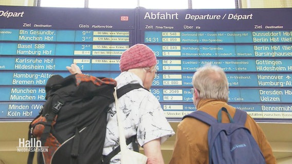 Zwei Reisende blicken auf eine Anzeigetafel in einem Bahnhof. © Screenshot 