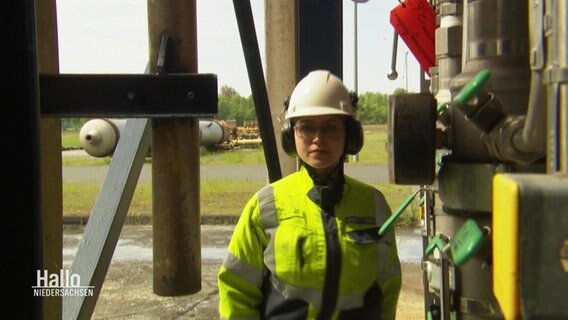 Eine Frau mit Schutzhelm und neongelber Arbeitskleidung geht an einer größeren Industrianlage vorbei. © Screenshot 