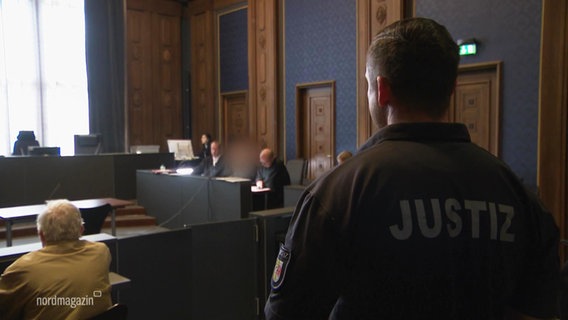 Blick in einen Gerichtssaal: Im Vordergrund steht ein Mitarbeiter der Justiz, im Hintergrund sitzt eine Angeklagter mit seinem Verteidiger auf der Anklagebank. © Screenshot 