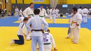 Mehrere Sportler in weißen Judo-Anzügen stehen auf Kampfsportmatten in einer Turnhalle. © Screenshot 