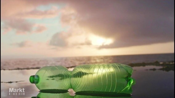 Eine grüne Plastik-Pfandflasche liegt in dem nassen Sand an einem Strand, im Hintergrund die untergehende Sonne über dem Meer. © Screenshot 