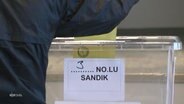 Eine Person mit schwarzer Jacke steckt einen gelben Umschlag durch eine gläserne Box mit der Aufschrift "3 No lu sandik". © Screenshot 