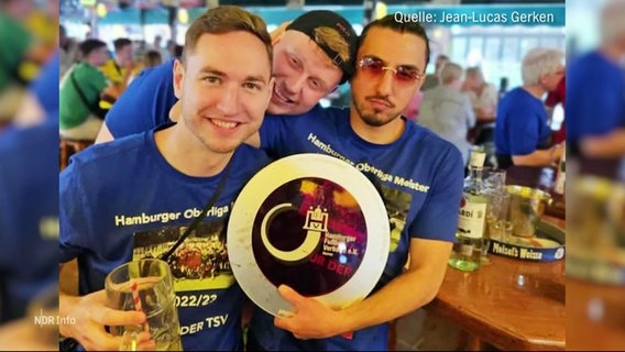Drei jüngere Männer in blauen Trikots halten an einer Bar einen kreisrunden Fußball-Pokal grinsend in die Kamera. © Screenshot 