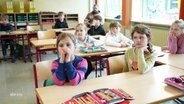 Grundschulkinder sitzen in einem Klassenraum in mehreren Reihen und blicken aufmerksam nach vorne. © Screenshot 
