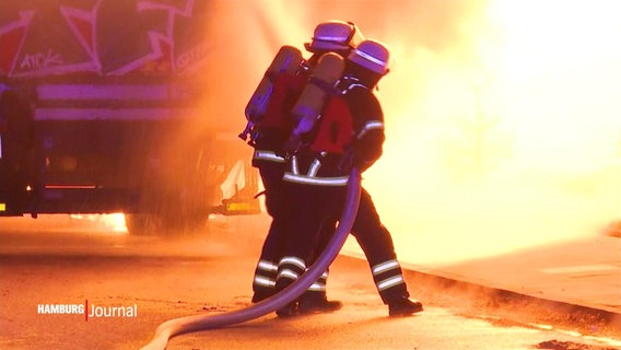 Zwei Einsatzkräfte der Feuerwehr halten gemeinsam einen Wasserschlauch, um gegen meterhohe Flammen anzukämpfen. © Screenshot 