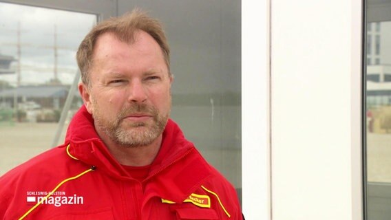 Thomas Becker von der DLRG Lübeck im Gespräch vor einer Rettungsschwimmer-Kabine am Strand © Screenshot 