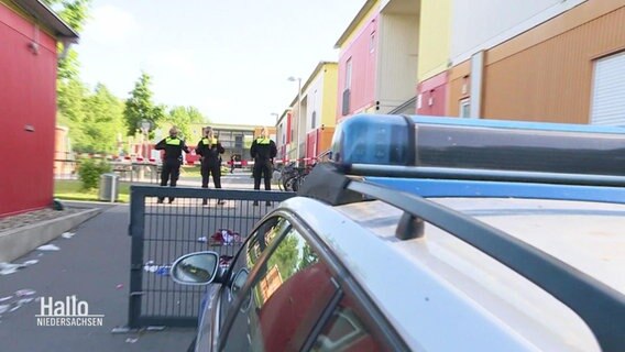 Blick auf das nicht eingeschaltete Blaulicht eines Polizeiautos, im Hintergrund stehen drei Einsatzkräfte der Polizei in einem Zufahrtsweg einer Wohnanlage. © Screenshot 