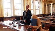 Der Angeklagte Michael Osterburg und sein Verteidiger im Gerichtssaal. Osterburg versteckt sein Gesicht hinter einer Mappe. © Screenshot 