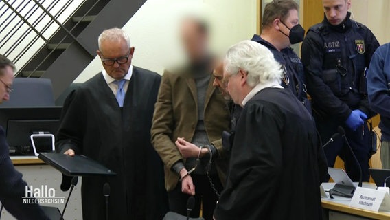 Ein Angeklagter mit unkenntlich gemachtem Gesicht steht mit Handschelle gefesselt neben seinen Verteidigern in einem Gerichtssaal. © Screenshot 