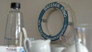 Im Vordergrund unscharf Geschirr und Gläser, im Hintergrund an der Wand ein weißer Rettungsring mit der blauen Aufschrift: "Weißer Ring - Wir helfen Kriminalitätsopfern". © Screenshot 
