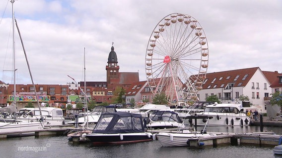 Blick auf einen kleineren Sportboothafen einer Kleinstadt an dem ein Riesenrad aufgebaut ist. © Screenshot 